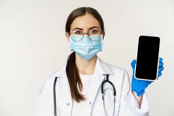 Online medical help concept. Woman doctor in glasses and face mask, showing mobile phone screen, app interface or website for patients, standing over white background - Fotoğraf, Görsel