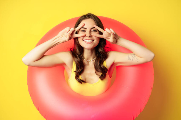 Portrait of attractive modern girl inside trendy pink beach swimming ring, showing peace v-sign and smiling happy, standing over yellow background - Photo, image