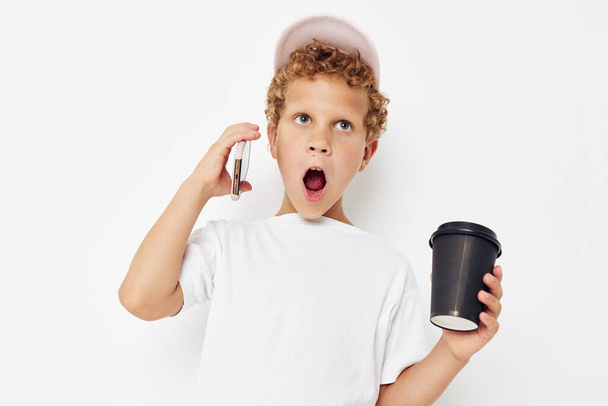 cute boy in a white t-shirt cap with a phone in a glass with a drink isolated background unaltered - Photo, Image