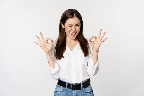 Smiling confident business woman, winking, showing okay ok sign, excellent, no problem gesture, standing over white background - Foto, immagini