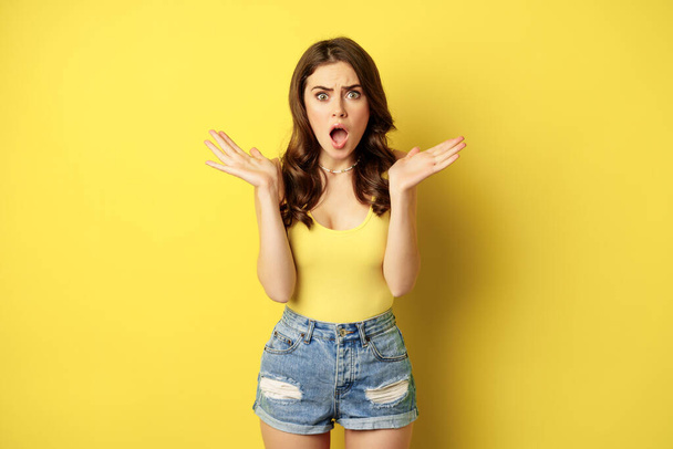 Shocked and frustrated woman gasping, looking overwhelmed and worried, concerned face, standing in summer tank top, yellow background - Foto, immagini