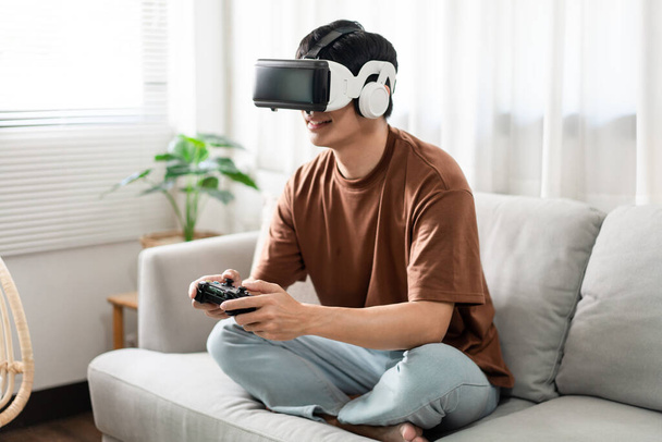 Concepto de tecnología Una persona que usa un auricular de realidad virtual y sostiene un juego de consola negro mientras está sentada en el sofá. - Foto, imagen