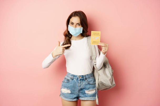 Portrait of woman in medical face mask travelling with covid international vaccination certification, holding backpack, standing over pink background - Foto, imagen