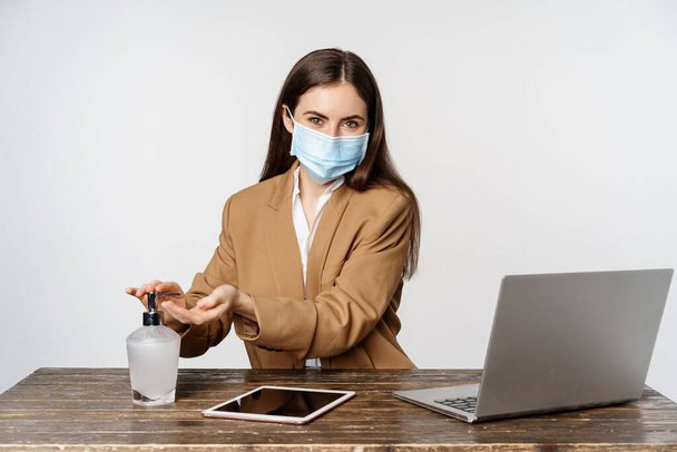 Workplace and covid-19 concept. Image of saleswoman, corporate woman in office cleansing hands with hand sanitizer and wearing face mask, white background - Photo, image