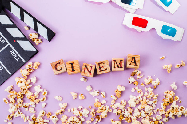 the word cinema, popcorn, clapperboard, 3D glasses and tickets top view flat lay on purple background - Foto, Imagem