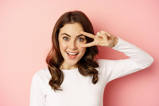 Close up portrait of coquettish young woman smiling, showing peace, v-sign gesture and posing happy, standing against pink background - Foto, Imagen