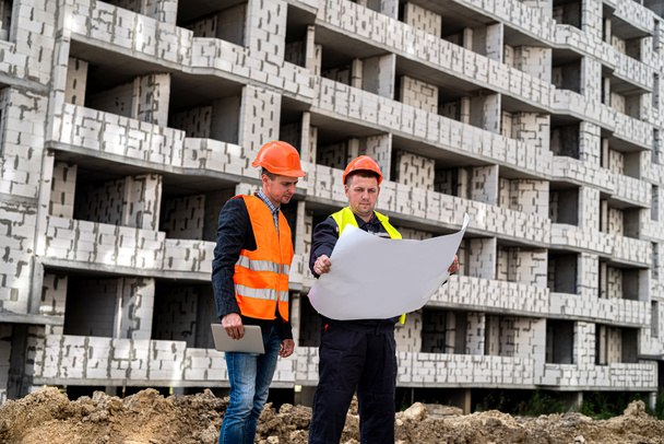 kräftige zwei Arbeiter in Helmuniformen mit Bauplan, die auf dem Bau von Häusern stehen. Baukonzept - Foto, Bild