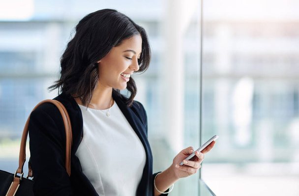 Her teams reporting good news today. Cropped shot of an attractive young businesswoman using a smartphone while standing in a modern office. - Foto, Imagem