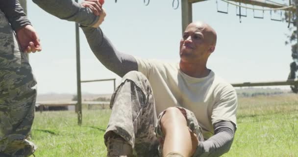 Female soldier helping diverse male colleague with injured leg at army obstacle course in the sun. healthy active lifestyle, cross training outdoors at boot camp. - Imágenes, Vídeo