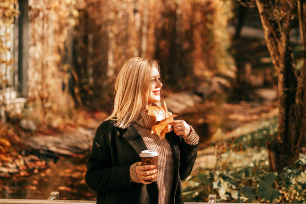 Bela paisagem de outono atrás sorrindo loira em casaco preto, iluminado pelo sol suave, ela caminha através do parque com buquês de folhas e xícara de café cappuccino. Estilo de vida. Vida no momento.  - Foto, Imagem