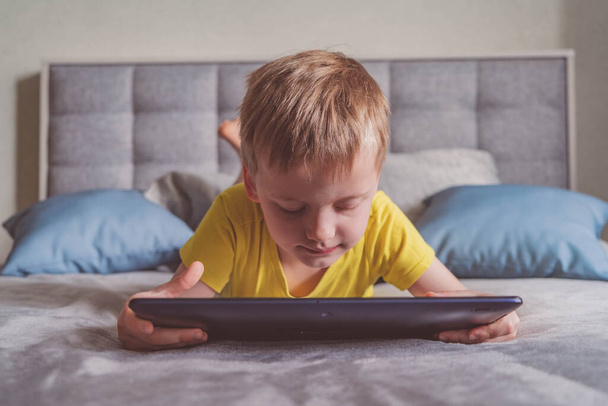 Distance education, learning. Boy is lying on the bed with a tablet in the room. Child with a gadget. Teenage child using tablet pc. - Photo, Image