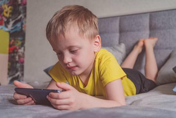 Educación a distancia, aprendizaje. El chico está acostado en la cama con un smartphone en la habitación. Niño con un gadget. Adolescente niño usando teléfono inteligente. - Foto, imagen