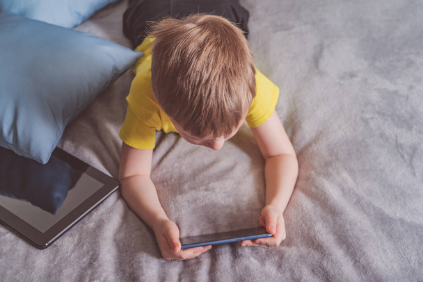 Distance education, learning. Boy is lying on the bed with a smartphone in the room. Child with a gadget. Teenage child using smartphone. - Photo, Image