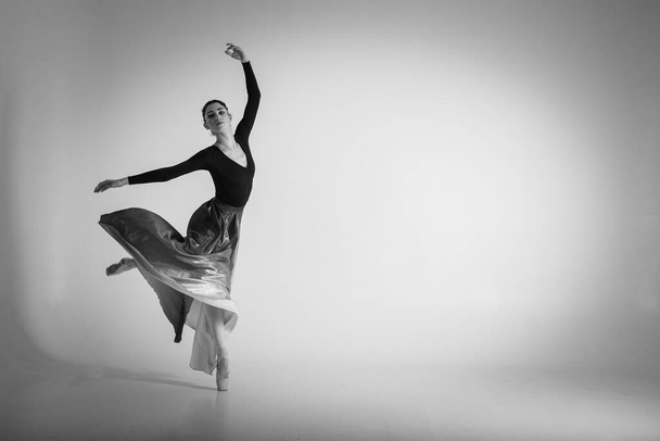 μπαλαρίνα. Μια νεαρή κομψή χορεύτρια μπαλέτου, ντυμένη με επαγγελματική ενδυμασία, παπούτσια και μια μπλε φούστα χωρίς βάρος, επιδεικνύει τις χορευτικές της ικανότητες. Η ομορφιά του κλασικού μπαλέτου. Ασπρόμαυρη φωτογραφία - Φωτογραφία, εικόνα