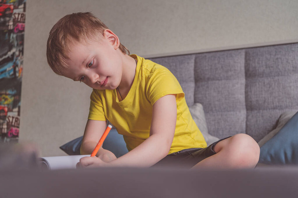 Молодий білий хлопчик малює в альбомі, пише на папері вдома, сидячи на ліжку
. - Фото, зображення