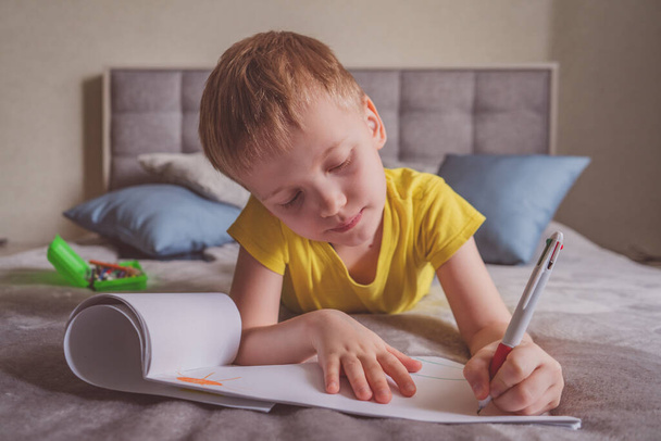 Young caucasian boy draws in an album, writes on paper at home sitting on bed. - Photo, image