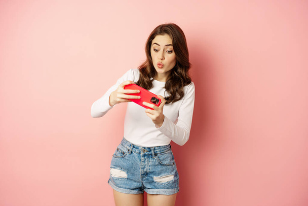 Gelukkig mooi meisje spelen mobiele video game, houden smartphone horizontaal, kijken op mobiele telefoon met opgewonden gezicht, roze achtergrond - Foto, afbeelding