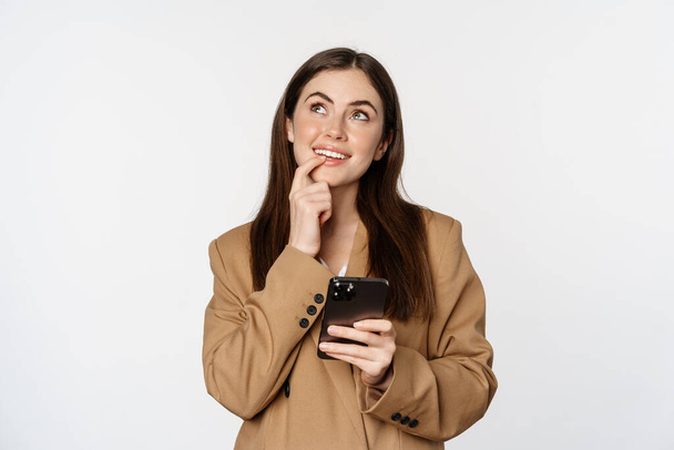 Image of businesswoman smiling, using mobile phone and thinking, making choice in smartphone app, standing against white background - Foto, Bild