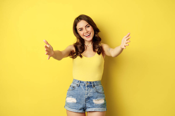Beautiful girl in tank top, reaching hands, stretching arms forward and smiling, welcome, hugging or greeting someone, standing over yellow background - Fotó, kép
