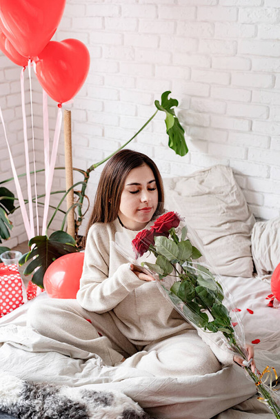 Valentine's day, Women's day. Young caucasian brunette woman sitting in the bed celebrating valentine day smelling red roses - Фото, изображение