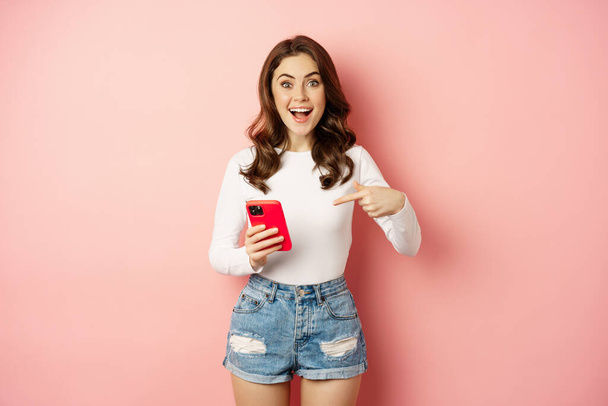 Happy brunette girl showing announcement, pointing finger at mobile phone, showing online promo, app news, standing against pink background - Foto, Bild