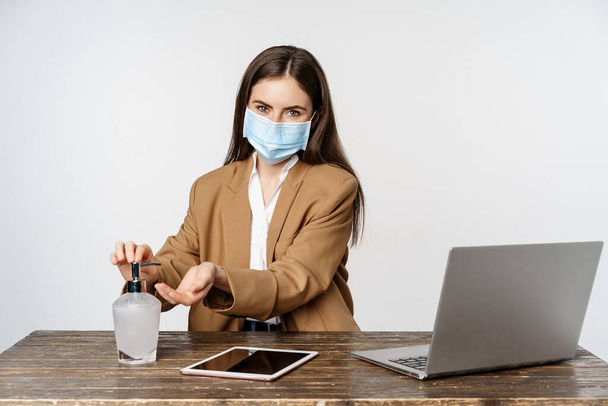 Podnikatelka na pracovišti nosí masku z covid-19, pracuje v kanceláři během pandemie, používá dezinfekci rukou k čištění rukou, pózuje na bílém pozadí - Fotografie, Obrázek