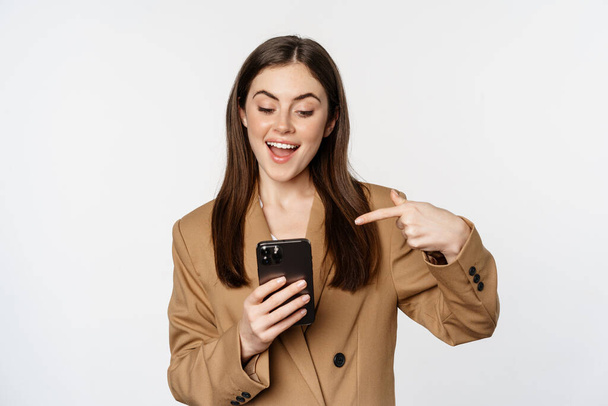 Nadšená prodavačka, obchodní žena ukazující prstem na mobilní telefon a usmívající se, ukazující na mobilu, stojící na bílém pozadí - Fotografie, Obrázek