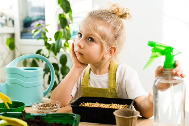 een klein blond meisje zit te broeden aan een houten tafel met haar wang op haar hand, groeiende micro-greens, water geven en spuiten met water uit een pulevizer. het begrip tuinieren - Foto, afbeelding