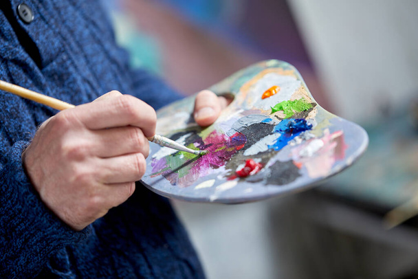 Closeup view of artist painting with palette, brush and acrylic colour paints in art studio indoors - Photo, image