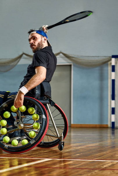 Νεαρός με σωματική αναπηρία παίζει τένις σε αναπηρικό καροτσάκι στο γήπεδο τένις. Μπάλες του τένις στερεωμένες σε τροχό - Φωτογραφία, εικόνα