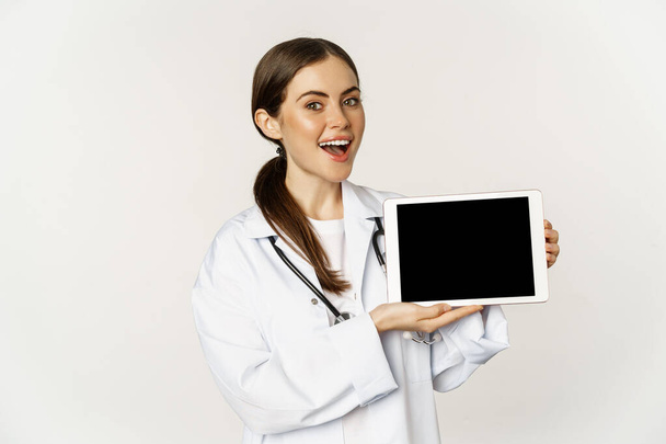 Online medical help, remote appointment. Smiling beautiful woman doctor showing digital tablet screen, demonstrating website logo, standing over white background - Zdjęcie, obraz