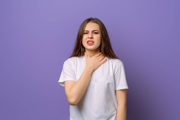 若いブルネットの女は病気で喉が痛い。不幸な女性の肖像、紫の背景に白いTシャツの上に立って - 写真・画像