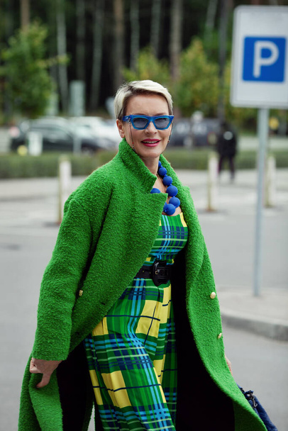 Portrait of fashionable adult woman wearing colorful bright accessories, sunglasses, green stylish coat. Bright fashion model with short blonde hair, street style - Фото, изображение