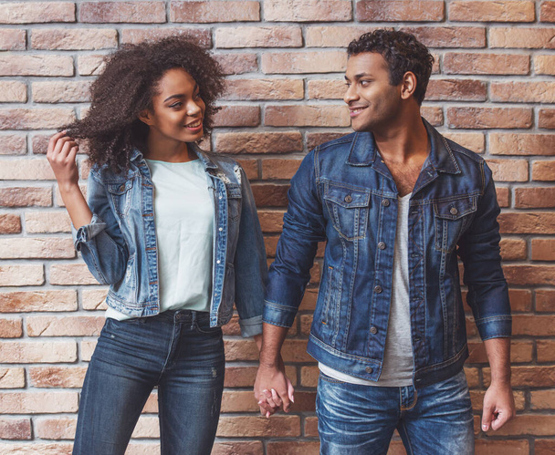 Jeune couple afro-américain attrayant se regardant, souriant et se tenant la main tout en se tenant contre un mur de briques
 - Photo, image