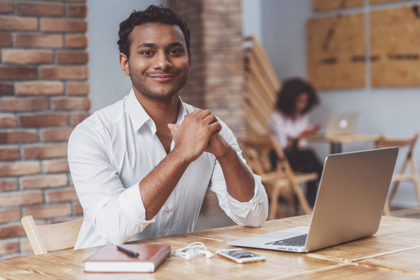 Молодий афро-американський бізнесмен у білій сорочці за допомогою ноутбука, дивиться в камеру і посміхається під час роботи в кафе. - Фото, зображення