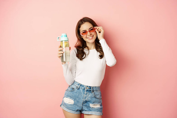 Stylish girl in spring outfit, wearing sunglasses, holding water bottle with lemon, healthy drink, laughing and smiling, standing over pink background - Фото, изображение
