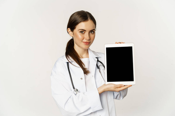 Image of woman doctor, female healthcare worker showing online medical website, digital tablet screen and smiling, standing in white coat over white background - Photo, Image