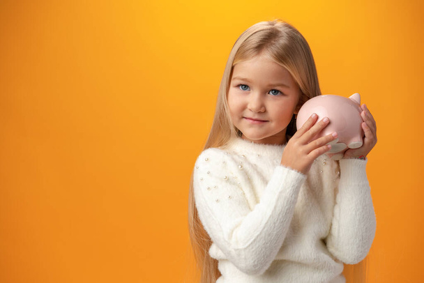Beautiful little girl with piggy bank on yellow background - 写真・画像