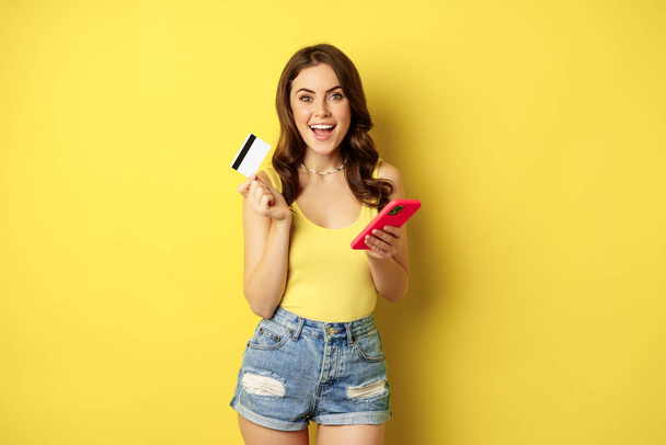 Online shopping. Stylish brunette woman holding smartphone and credit card, paying in app, using mobile phone application, buying smth, standing over yellow background - Foto, Imagen