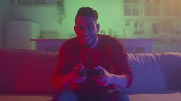 Tv pov portrait of emotional middle eastern guy playing video games with joystick, sitting on sofa at home in evening - 映像、動画
