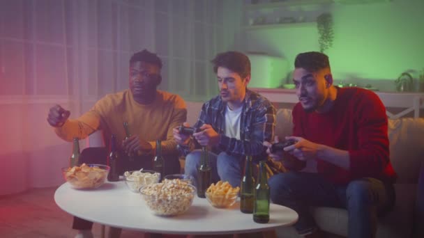 Vidám, fiatal, változatos férfi barátok videojátékoznak joystickokkal, este otthon pihennek sörrel és snackkel - Felvétel, videó
