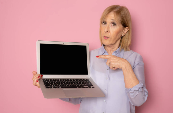 Mujer mayor que muestra la pantalla del ordenador portátil de pie aislado sobre fondo rosa - Foto, imagen