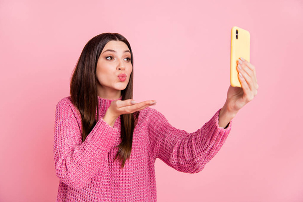 Фото блестящей милой молодой женщины в трикотажном свитере с селфи, посылающей руку поцелуй современные устройства изолированный розовый цвет фона. - Фото, изображение