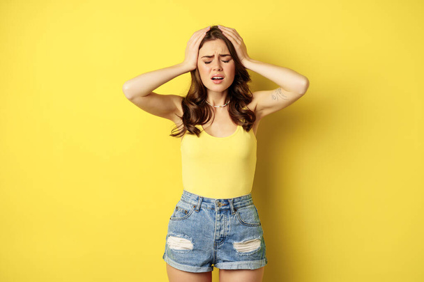 Troubled and shocked woman holding hands on head, panicking, feeling distressed and anxious, having problem, standing over yellow background - Foto, Imagen