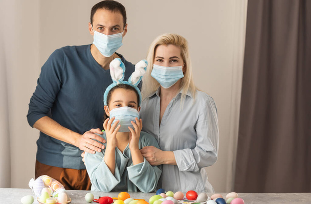 Pap, mam en kind aan tafel met medische maskers schilderen paaseieren voor de feestdag. Gelukkige Pasen familie in quarantaine geplaatst coronavirus. Thuis gelukkig. - Foto, afbeelding