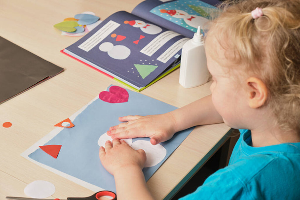 a preschooler girl in a blue T-shirt makes an applique while sitting at the table, a paper snowman hauled on colored paper, children's creativity in winter, atopic dermatitis on the hands of a child - Photo, image