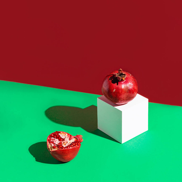 Kreativní složení z čerstvých granátových jablek na zeleném a červeném pozadí s bílou kostkou a stíny. Minimální styl. Koncept občerstvení. Téma zdravé složky potravin. - Fotografie, Obrázek