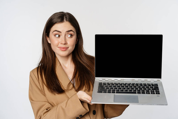 Young woman cringe and look skeptical, showing laptop screen, standing over white background - Foto, immagini