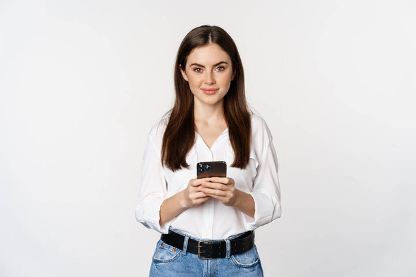 Молода жінка зі смартфоном, посміхаючись і дивлячись на камеру, використовуючи мобільний додаток, мобільні технології та концепцію інтернет-магазинів, білий фон
 - Фото, зображення