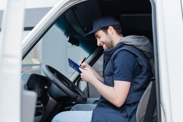 Side view of smiling courier writing on clipboard near steering wheel in car  - 写真・画像
